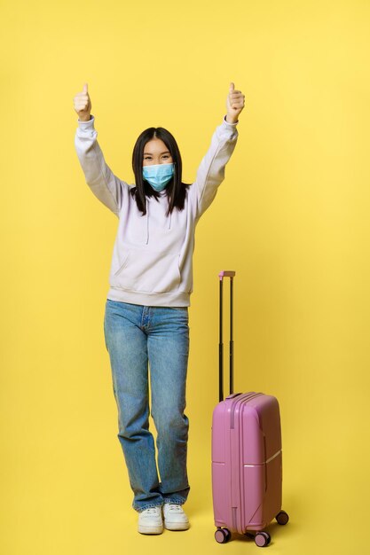 Снимок в полный рост счастливой азиатской туристки в отпуске в медицинской маске, показывает палец вверх и ...