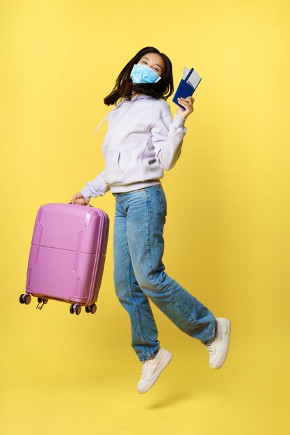 Full length shot happy asian woman jumping with suitcase and tickets on tour, holding passport, going on vacation, standing over yellow background