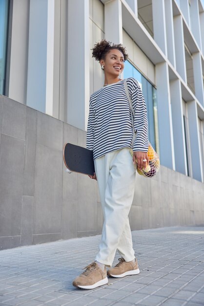 Full length shot of happy curly haired woman dressed in striped jumper white trousers and sneakers carries net bag and longboard stands near urban building strolls outdoors has glad expression