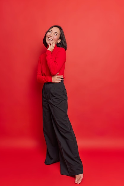 Full length shot of happy dreamy brunette young Asian woman has positive expression wears turtleneck black loose trousers stands against vivid red wall thinks about something very pleasant