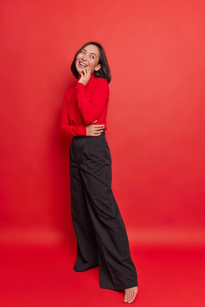 Full length shot of happy dreamy brunette young Asian woman has positive expression wears turtleneck black loose trousers stands against vivid red wall thinks about something very pleasant