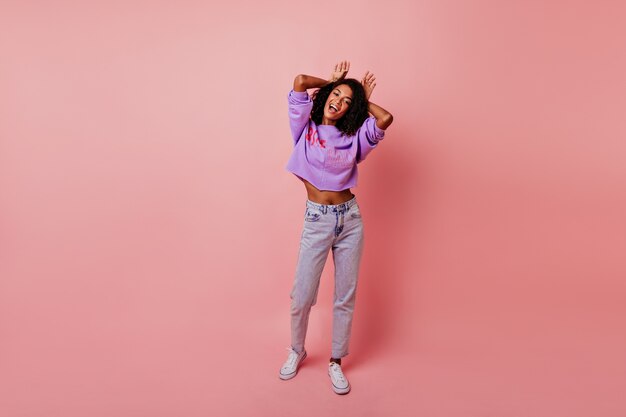 Full-length shot of inspired black girl joking during portraitshoot. Carefree curly woman making funny faces on pink.