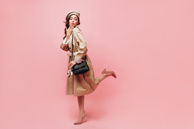 Full-length shot of lady in midi trench coat coquettishly lifting leg and blowing kiss on pink background.