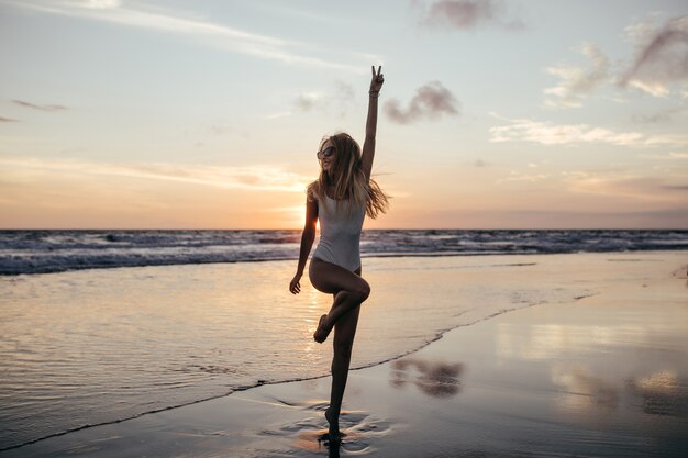바다 해안에서 한쪽 다리에 서있는 사랑스러운 슬림 소녀의 전체 길이 샷.