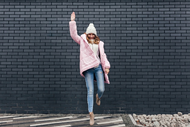 Full-length shot of magnificent girl in jeans and winter accessories. Outdoor portrait of good-looking blonde woman dancing on urban street.