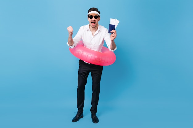 Full-length shot of man enjoying vacation trip. Guy in business suit and sunglasses holding documents, tickets and pink inflatable circle.