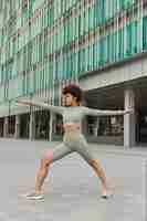 Free photo full length shot of motivated curly haired sporty woman stretches arms exercises outdoors dressed in activewear poses near modern urban building focused into distance workout and sport concept