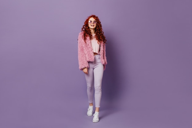 Full-length shot of naughty red-haired girl in round glasses, white jeans and pink coat on purple space.