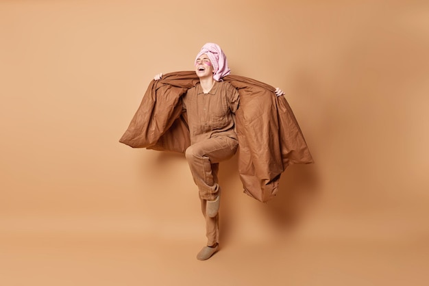 Full length shot of positive young woman dressed in pajama stands on one leg foolishhes around dances with blanket has wrapped towel on head isolated over beige background applies patches under eyes