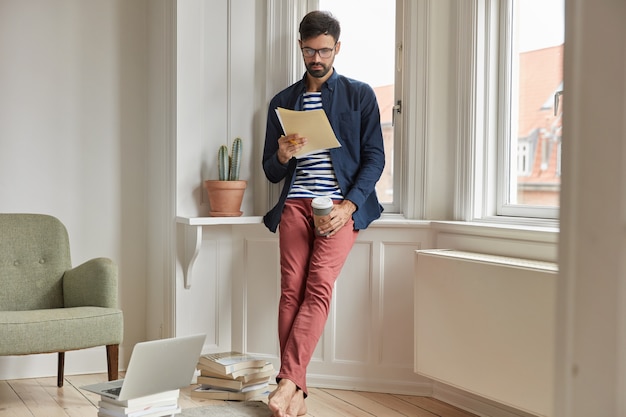 Full length shot of serious unshaven man or entrepreneur studies invoice, does paper work at home