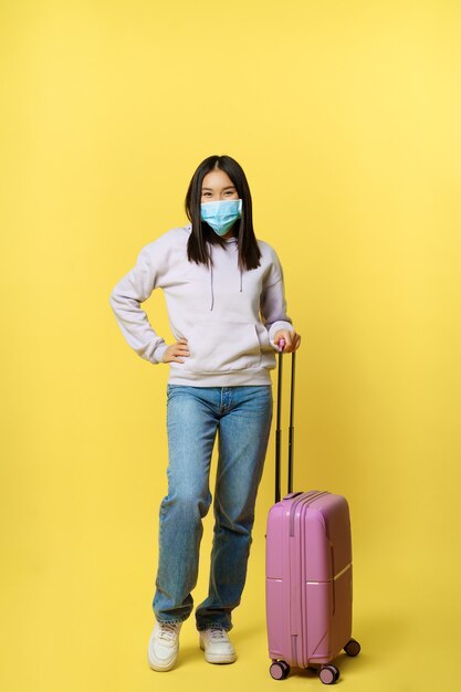 전체 길이 사진은 의료용 얼굴 마스크를 쓰고 여행가방을 들고 포즈를 취하고 휴가를 가고 노란색 배경 위에 서 있는 한국 여성 관광객을 웃고 있습니다.
