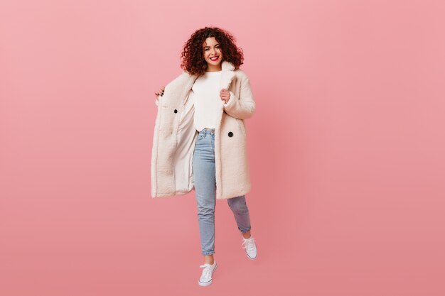 Full-length shot of smiling lady dressed in eco-coat, white sweater and denim pants on pink space.