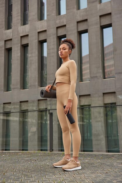 Full length shot of sporty young woman dressed in tracksuit carries rolled karemat holds bottle of water poses agains modern building going to have cardio training prepares for morning workout