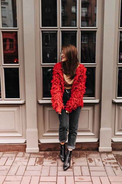 Full-length shot of stylish woman in black jeans and red cardigan, leaning on white window in street.