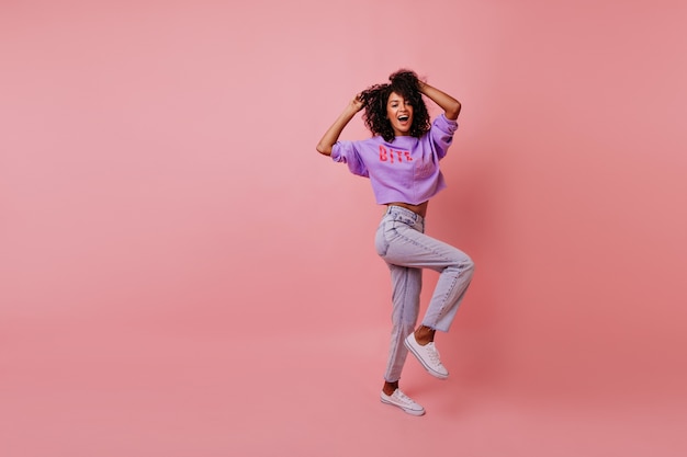 Full-length shot of stylish woman dancing on rosy. Appealing female model in jeans fooling around in studio.