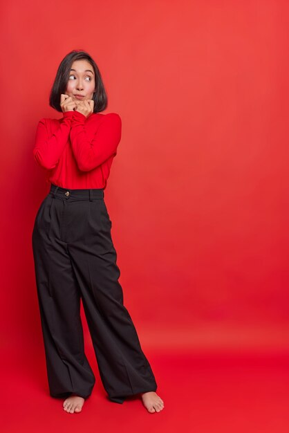 Full length shot of thoughtful Asian woman with dark hair keeps hands on chin looks away at something interesting wears turtleneck black loose trousers stands bare feet indoor against red wall