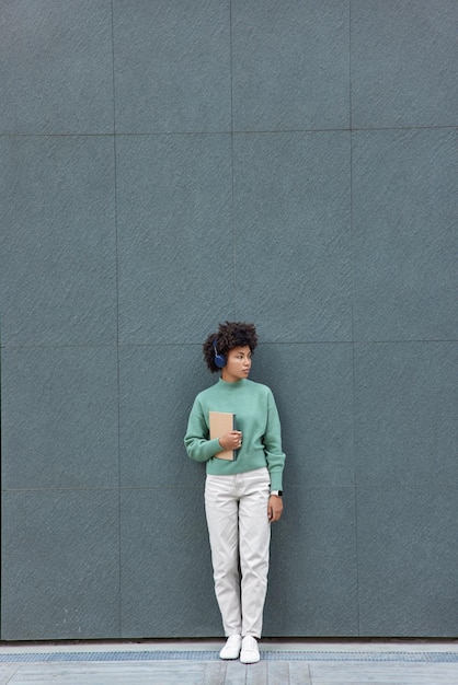 Full length shot of thoughtful female student holds notepad returns from lectures listens autodidact concentrated away wears casual jumper trousers and sneakers poses against blank grey wall