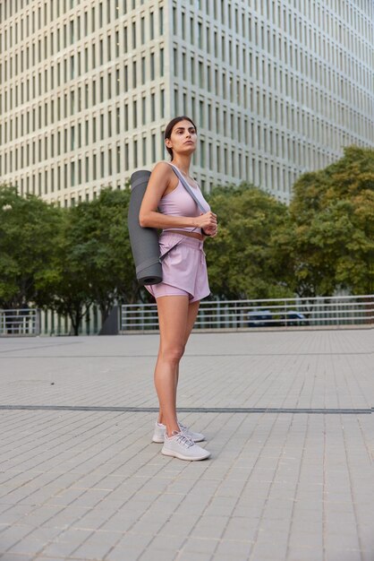 Full length shot of thoughtful sporswoman or yoga instructor has good shape dressed in cropped top shorts and sneakers carries fitness mat poses in city against skyscrapers and green tress around.