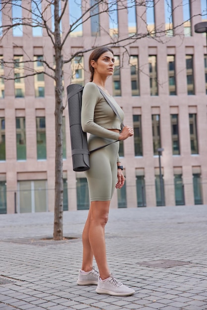 Full length shot of thoughtful sporty woman dressed in activewears and sneakers carries rolled fitness mat going to have yoga practice outdoors poses against modern city building. Aerobics concept