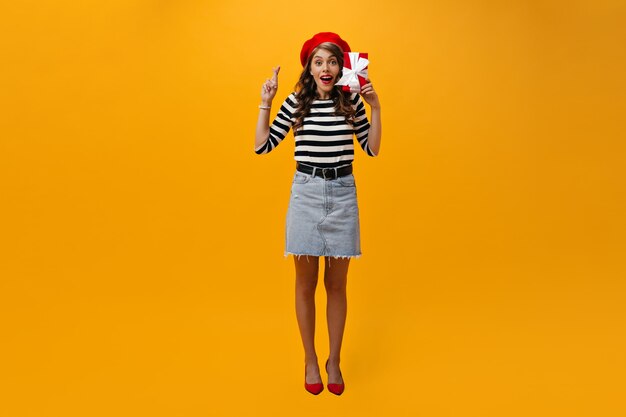 Full length shot of woman crossing fingers and holding red gift box. Happy girl in red beret and striped shirt looking into camera.