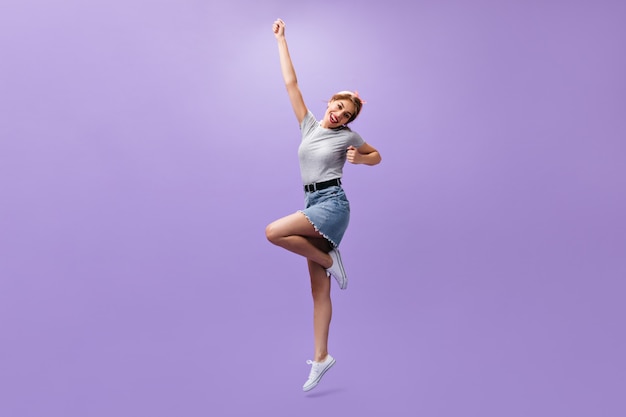 Full length shot of woman in denim skirt on purple background. Slim cool girl with pink bandana and trendy hairstyle in summer clothes jumping.
