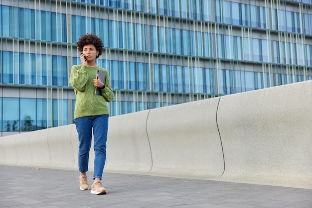 Full length shot of young curly haired woman has telephone phone and walks outdoors against modern city building uses modern gadgets communicates via cellular during spare time. people and lifestyle
