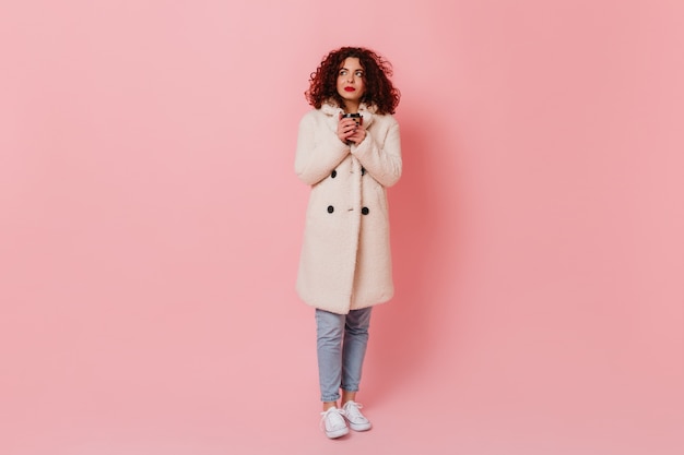 Full-length shot of young curly woman with red lips wearing white coat and jeans holding glass of tea on pink space.