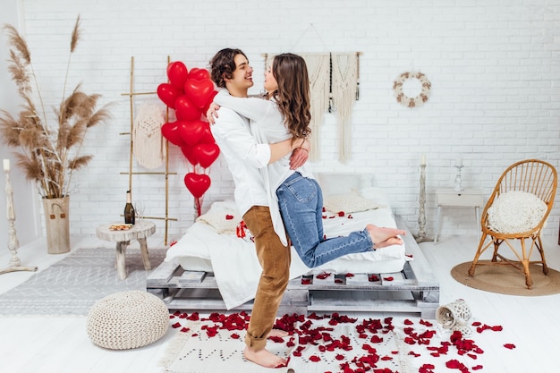 Молодой человек держит свою девушку на руках в полный рост в комнате, украшенной лепестками роз, на день святого валентина