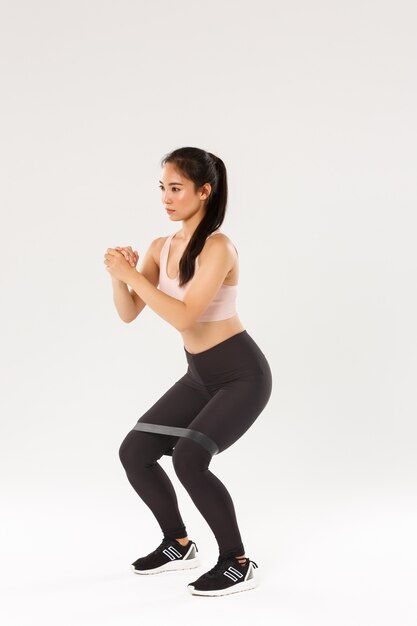 Full length side view of focused slim asian girl doing fitness training, female athlete clasp hands together and perform squats exercises with stretching resistance band, workout equipment.