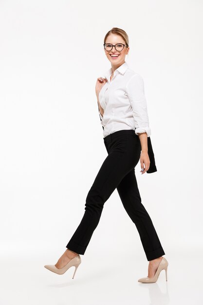Full length side view image of smiling blonde business woman in eyeglasses walking in studio and  over white wall