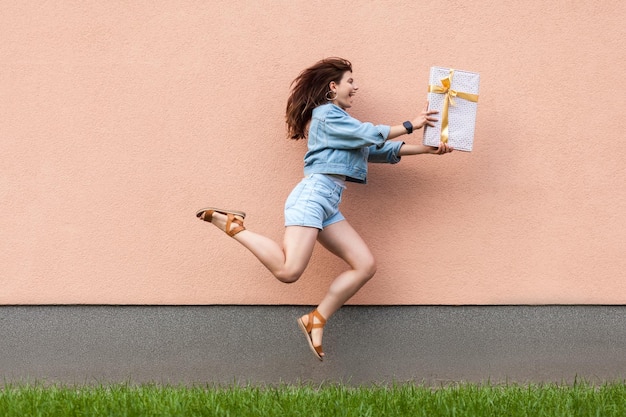 여름에 캐주얼 청바지 데님 스타일을 입은 행복한 흥분된 아름다운 여성의 전체 길이 측면 전망은 모래갈색 벽 근처에 서서 선물 상자를 들고 점프합니다. 쇼핑, 축하 또는 배달 개념.