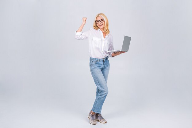 白​灰色​の​背景​で​隔離​の​ラップ​トップ​pc​コンピューター​で​作業して​立っている​陽気な​高齢者​成熟した​シニア​ブロンド​女性​女性​40​代​50​代​の​笑い​の​完全な​長さ​の​側面図​。