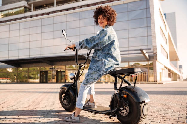 Full-length side view of smiling curly woman sitting on motorbike