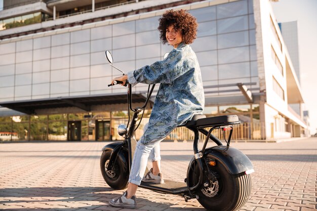 Full-length side view of smiling curly woman sitting on motorbike