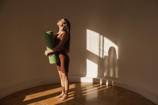 Бесплатное фото Стройная женщина в полный рост с фитнес-телом в верхних леггинсах, держащая коврик для упражнений, стоя в помещении девушка с темными длинными волосами собирается на тренировку домашняя спортивная концепция