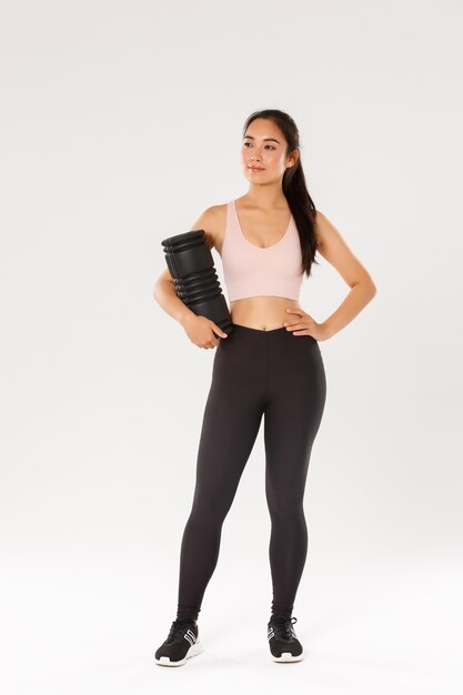 Full length of smiling confident and motivated asian female gym coach, fitness girl in sportswear, looking around pleased, holding foam roller to use after workout, standing with training equipment.