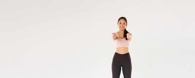 Full length of smiling goodlooking and slim asian fitness coach female athlete in activewear showing