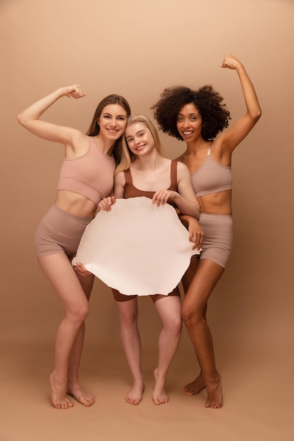 Foto gratuita tre giovani donne interrazziali sorridenti a tutta lunghezza che tengono un cartello bianco su sfondo beige concetto di emozioni positive