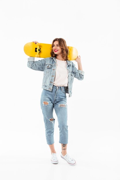 Full length Smiling woman in denim clothes holding skateboar and looking away over white wall