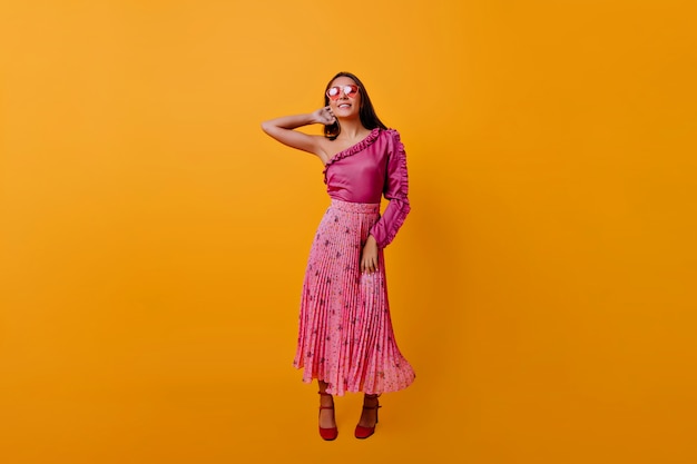 Снимок в полный рост в оранжевой комнате на изолированной стене. Хорошо сделанная женщина в розовом топе и макси-юбке кокетливо трогает шею