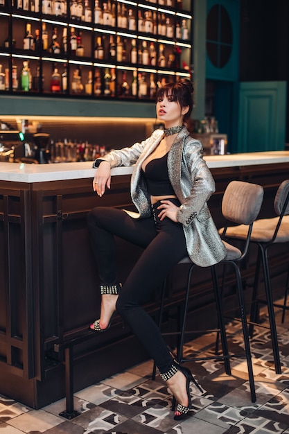 Full length stock photo of an extremely trendy luxurious brunette model in crop top, silver sparkling jacket, black trousers and high heels. Model in trendy outfit sitting on bar stool in club or bar.