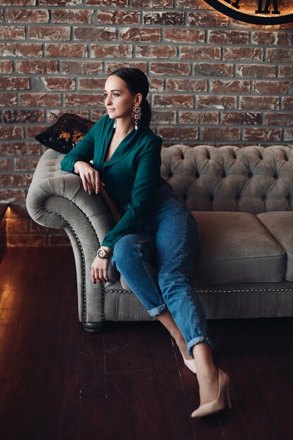 Full length stock photo of gorgeous brunette woman in casual elegant clothes and high heels relaxing on classic sofa with cushions and looking away. The room is in loft style.