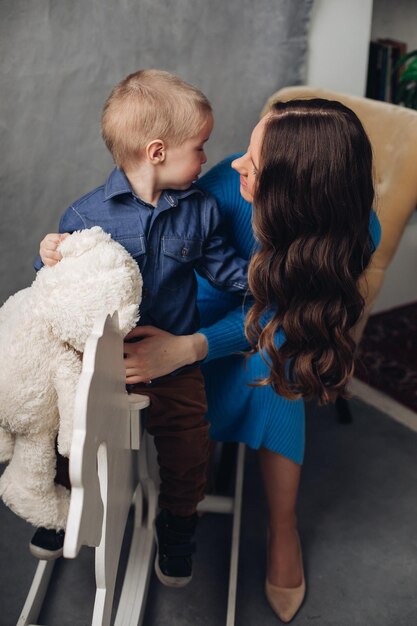 Full length studio portrait of gorgeous elegant brunette mom in denim dress with lovely laughing son on her hands They smiling at camera