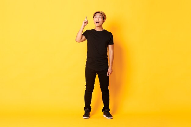 Full-length of thoughtful handsome asian man, raising finger up, have good idea, standing yellow wall