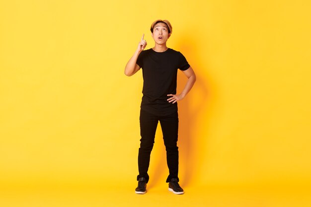 Full-length of thoughtful, intrigued attractive blond asian man raising finger, pointing up and looking amused, yellow wall