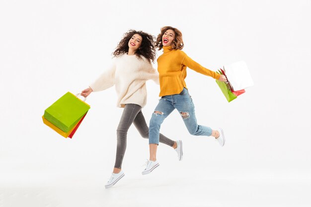 Full length Two joyful girls in sweaters running together with packages and looking away over white wall