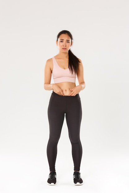 Free photo full length of upset and gloomy asian girl in fitness clothing, showing fat on belly, complaining on body, frowning and looking disappointed, starting workout, trying lose weight, white background.