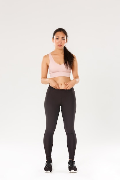 Full length of upset and gloomy asian girl in fitness clothing, showing fat on belly, complaining on body, frowning and looking disappointed, starting workout, trying lose weight, white background.