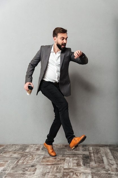 Full-length uptight man with takeaway coffee looking at watch being late, and running along gray