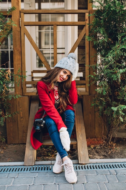 Полнометражная вертикальная девушка брюнет с длинными волосами в красном пальто и связанной шляпе, сидя на деревянной лестнице на открытом воздухе. Она носит теплые белые перчатки и улыбается.